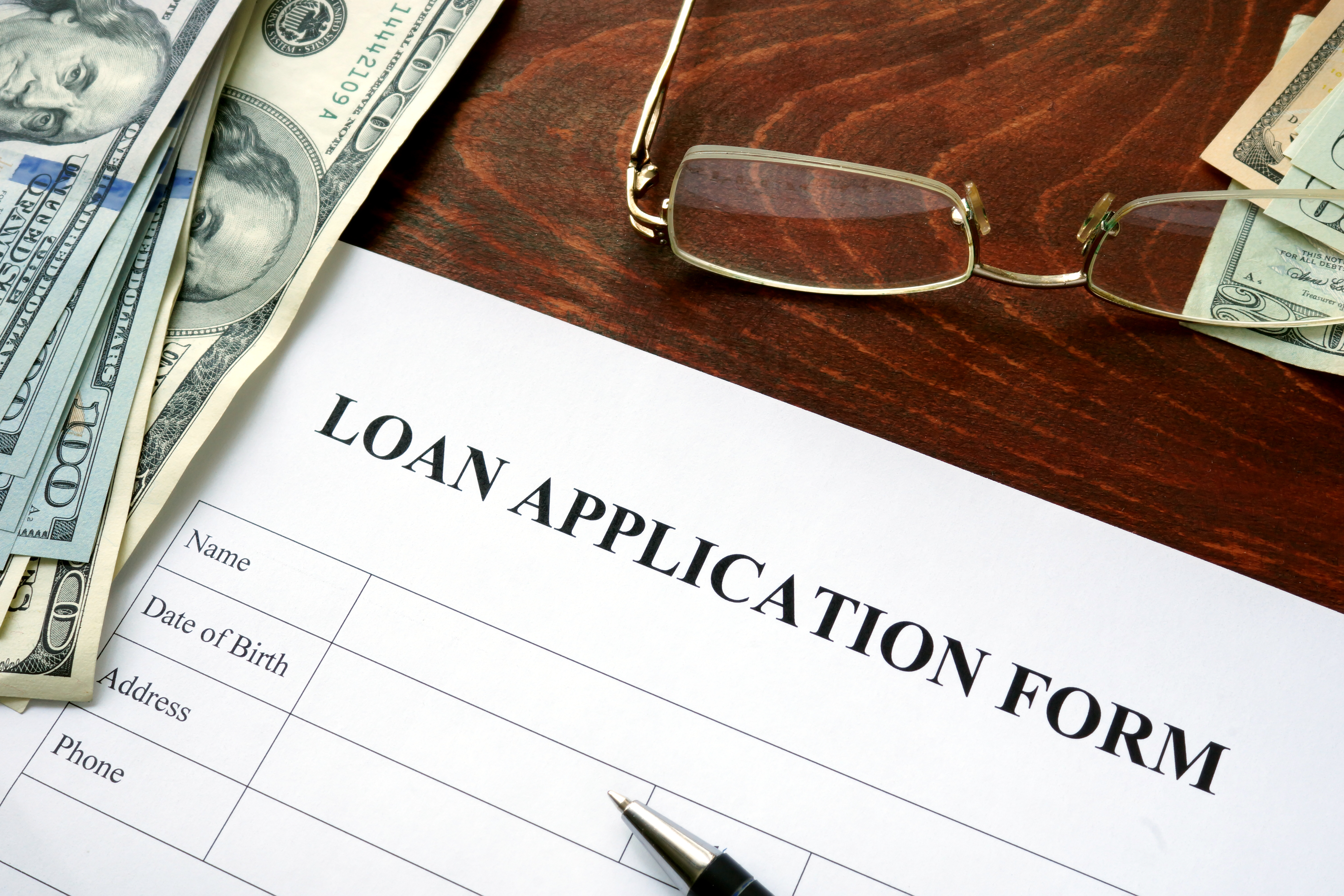 A loan application form, several one-hundred dollar bills, a pair of glasses, and a pen arranged on a desktop.